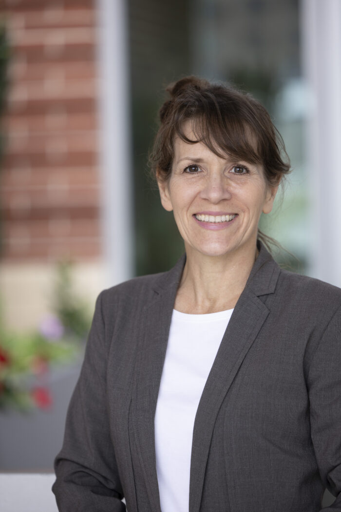 woman with brown hair, white shirt, and gray blazer smiling at camera